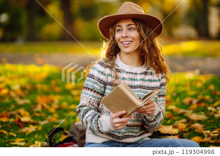 Beautiful curly woman in hat on mat with book in autumn park. Relaxation, solitude with nature. 119304998