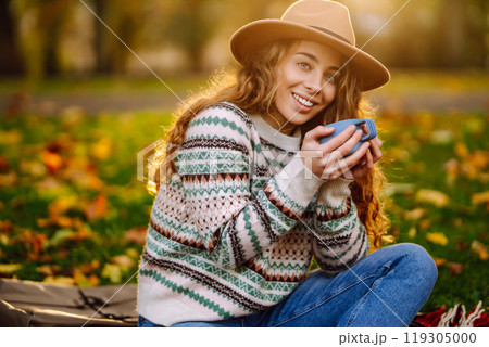 Curly woman with thermos in clearing among fallen leaves in autumn park, enjoying hot drink. Curly woman with thermos in clearing among fallen leaves in autumn park, enjoying hot drink. 119305000