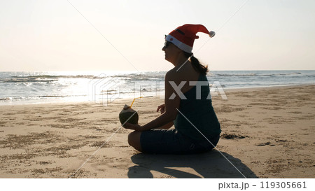 Silhouette of a young woman in a festive Christmas hat relaxes by the seashore, sipping refreshing coconut water while enjoying the winter holidays 119305661