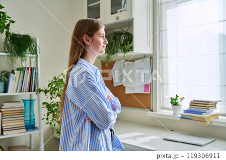 Smiling confident young woman looking out the window, in home interior Smiling confident young woman looking out the window, in home interior 119306911