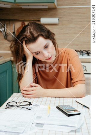 sad woman pays bills online, sits in kitchen with receipts and calculator. Online banking  119307141