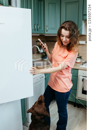 happy European woman looks into refrigerator and orders food using lady's smartphone. happy European woman looks into refrigerator and orders food using lady's smartphone. 119307154