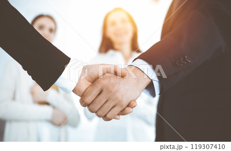Businessman and woman shaking hands with colleagues at the background. Handshake at meeting in sunny office Businessman and woman shaking hands with colleagues at the background. Handshake at meeting in sunny office 119307410