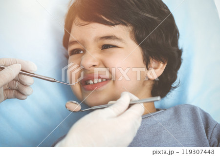 Cute arab boy sitting at dental chair with open mouth during oral checking up with doctor. Visiting sunny dentist office Cute arab boy sitting at dental chair with open mouth during oral checking up with doctor. Visiting sunny dentist office 119307448
