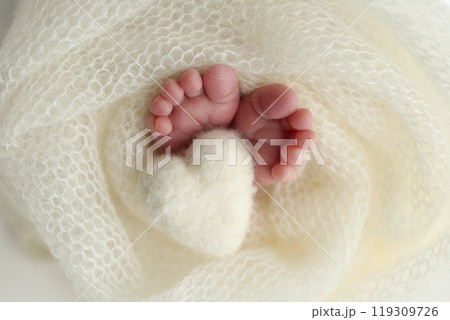 The tiny foot of a newborn baby. Soft feet of a new born in a white wool blanket. Close up of toes, heels and feet of a newborn. Knitted white heart in the legs of a baby. Macro photography.  119309726