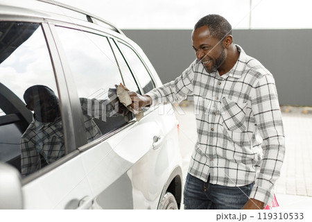 Man washing luxury car on a car wash with a rag. Black man wiping his car. Man wearing plaid shirt, smiling. Man washing luxury car on a car wash with a rag. Black man wiping his car. Man wearing plaid shirt, smiling. 119310533