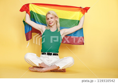 Young blonde man with make up on his face and rainbow flag posing for a photo. Man wearing fashionable green top and has red lips. Man isolated on yellow background. Young blonde man with make up on his face and rainbow flag posing for a photo. Man wearing fashionable green top and has red lips. Man isolated on yellow background. 119310542