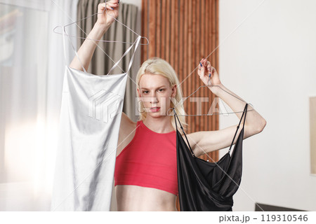 Man with make up on his face standing in a bedroom in his flat. Man wearing fashionable pink top, white trousers. Man choosing what to wear: black or white dress. 119310546