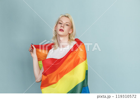 Young blonde man with make up on his face and rainbow flag posing for a photo. Man wearing fashionable white t-shirt and has red lips. Man isolated on blue background. 119310572