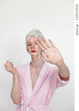 Young man with make up on his face doing manicure. Man wearing pink dressing gown and has a towel on his head. Man isolated on white background. 119310582