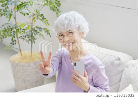 スマートフォンを持ってピースサインするシニア女性　リビングルーム 119313032