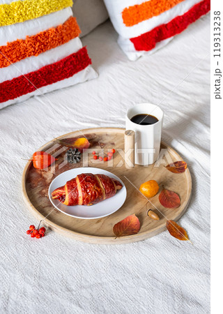 Cozy breakfast tray featuring coffee and croissant on bed, with autumn leaves around. 119313238