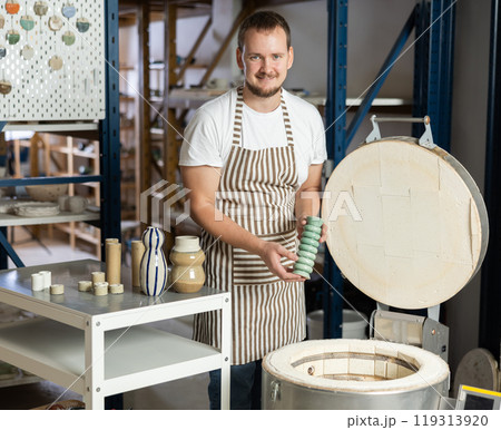 European guy in apron takes ready-made pottery made of clay out of oven 119313920
