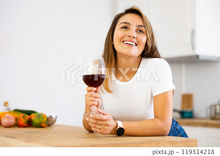 Cheerful young Latina drinking wine in home kitchen 119314218