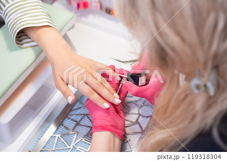 Nail technician in pink gloves skillfully applies nude gel polish to a clients hand while ensuring perfect nails. 119318304