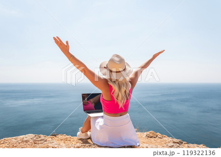 A woman is sitting on a rock by the ocean with a laptop open in front of her. She is wearing a pink top and a straw hat. Concept of relaxation and leisure. A woman is sitting on a rock by the ocean with a laptop open in front of her. She is wearing a pink top and a straw hat. Concept of relaxation and leisure. 119322136