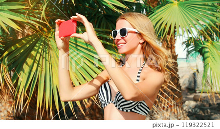 Happy smiling young woman taking selfie with smartphone on the beach on palm tree background 119322521