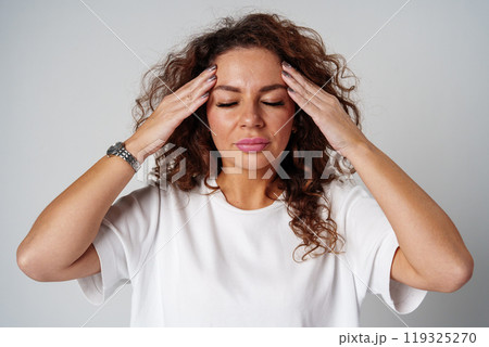Woman with curly hair holds her head in discomfort, expressing concern or stress in a neutral backdrop 119325270