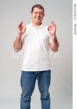 A man in a white shirt poses with a cheerful expression showing OK hand gestures in a well-lit indoor setting A man in a white shirt poses with a cheerful expression showing OK hand gestures in a well-lit indoor setting 119325288