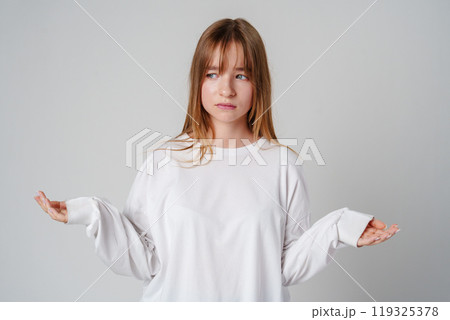 A young girl wearing a white long-sleeve shirt expressing confusion in a minimalistic gray studio setting 119325378