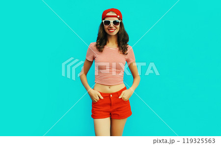 Summer portrait of happy smiling young woman wearing baseball cap, red shorts on blue background 119325563