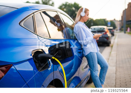 A blonde woman relaxes with headphones standing next to her electric car, charging by the roadside in the evening. 119325564