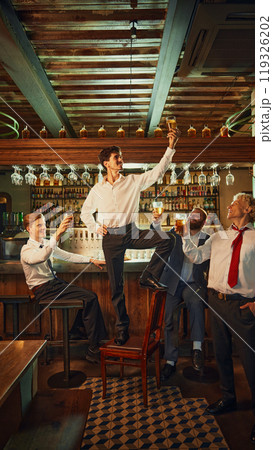 Businessman raises glass triumphantly in bar, surrounded by colleagues who cheer him on, creating celebratory atmosphere. Businessman raises glass triumphantly in bar, surrounded by colleagues who cheer him on, creating celebratory atmosphere. 119326202