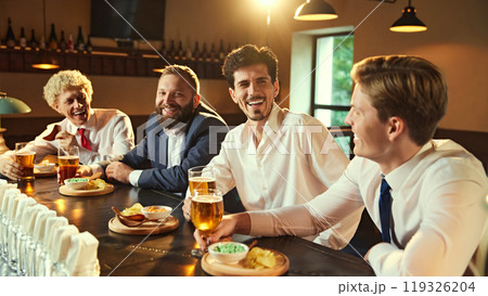 Men share stories and laughs at bar, each with beer in hand, creating perfect moment of connection and shared experience. 119326204