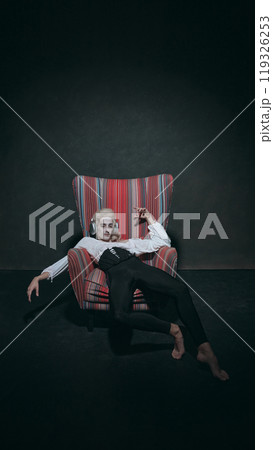 Reclining in colorful armchair, man in historical attire listening music in modern headphones, symbolizing relaxation against vintage background. 119326253