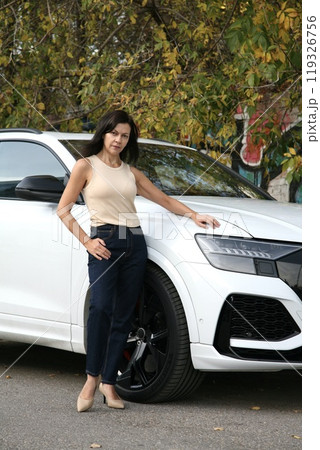 Beautiful brunette woman with long straight hair standing and posing near white sports car against autumn street background 119326756
