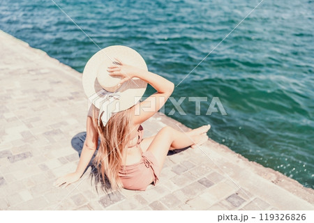 A woman in a swimsuit sits with her back holding a hat, looks at the ocean, sunny day, relaxes. A woman in a swimsuit sits with her back holding a hat, looks at the ocean, sunny day, relaxes. 119326826
