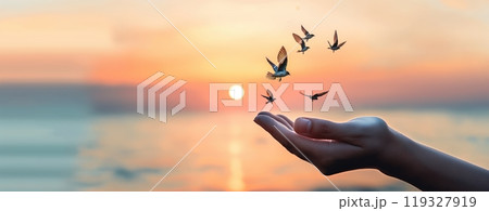 A gentle hand extends towards the horizon, where birds are taking flight against a vibrant sunset over the calm sea. 119327919