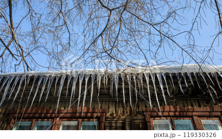 View from bottom to row of spring icicles hanging down from snow covered roof of old wooden house. 119330326