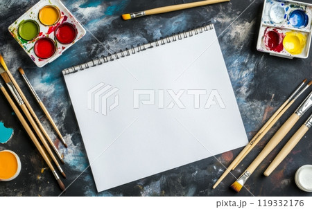 A blank sketchpad surrounded by vibrant watercolor paints, brushes, and palette on a textured background, representing an artist's creative workspace 119332176