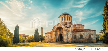 Picturesque Byzantine style church with domed roof and arched windows, surrounded by cypress trees and bathed in warm evening light 119332188