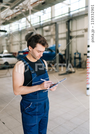 Vertical shot of focused auto mechanic man checking car with clipboard in garage car service. Serious technician male repairing vehicle at garage, writing filling checklist in auto repair shop. Vertical shot of focused auto mechanic man checking car with clipboard in garage car service. Serious technician male repairing vehicle at garage, writing filling checklist in auto repair shop. 119332901