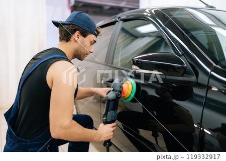 Rear view of muscular mechanic worker polishing door of luxury car in auto repair shop using with orbital polisher. Expert car paint technician using car polisher to polish car with refinement. Rear view of muscular mechanic worker polishing door of luxury car in auto repair shop using with orbital polisher. Expert car paint technician using car polisher to polish car with refinement. 119332917