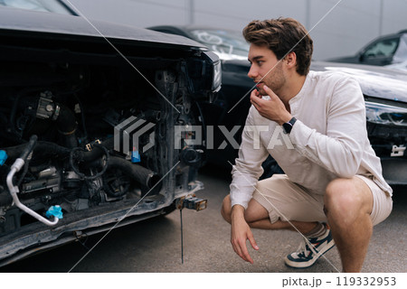 Stressed car owner male squatting and inspecting damage to car after accident. Frustrated driver looking damaged car. Concept of buying car accident insurance. 119332953