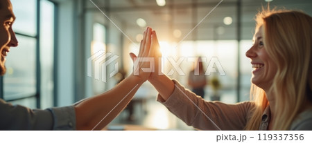 Two women share a high five in a bright indoor setting, celebrating success or teamwork. The image represents positivity, collaboration, and achievement. 119337356