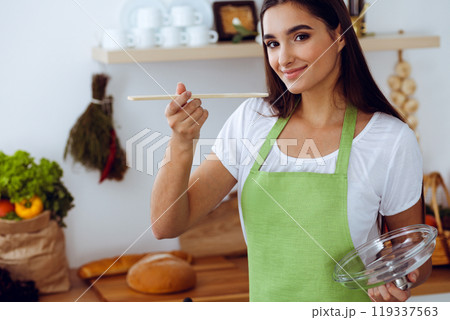 An attractive young dark-haired woman tasting ready hot meal with a wooden spoon while standing and smiling near the kitchen stove. Cooking and householding concepts An attractive young dark-haired woman tasting ready hot meal with a wooden spoon while standing and smiling near the kitchen stove. Cooking and householding concepts 119337563