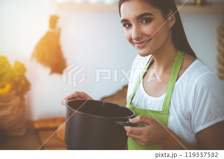 An attractive young dark-haired woman tasting ready hot meal with a wooden spoon while standing and smiling in sunny kitchen. Cooking and householding concepts An attractive young dark-haired woman tasting ready hot meal with a wooden spoon while standing and smiling in sunny kitchen. Cooking and householding concepts 119337582