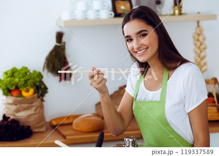 An attractive young dark-haired woman tasting ready hot meal with a wooden spoon while standing and smiling near the kitchen stove. Cooking and householding concepts An attractive young dark-haired woman tasting ready hot meal with a wooden spoon while standing and smiling near the kitchen stove. Cooking and householding concepts 119337589