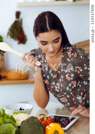 An attractive young dark-haired woman tastes a new recipe for a delicious salad mix while sitting at the table in the kitchen. Tablet pc is the best cookbook 119337606