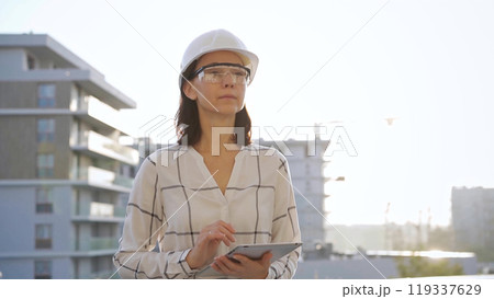 Woman construction engineer wearing white checked blouse and hard hat is using tablet computer while inspecting a building site at sunset, front view. Architecture and engineering concepts 119337629
