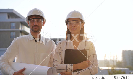 Man and woman architects, wearing safety helmets, are standing near each other and looking at the camera at a construction site during sunrise or sunset. Architecture and engineering concept Man and woman architects, wearing safety helmets, are standing near each other and looking at the camera at a construction site during sunrise or sunset. Architecture and engineering concept 119337630