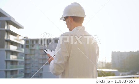 Man construction engineer wearing white shirt and hard hat is making notes on a clipboard while inspecting a building site at sunset, back view. Architecture and engineering Man construction engineer wearing white shirt and hard hat is making notes on a clipboard while inspecting a building site at sunset, back view. Architecture and engineering 119337657