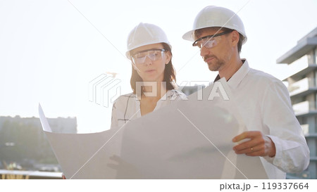 Two architects, wearing safety helmets and glasses, are examining blueprints at a construction site during sunrise or sunset, front view. Architecture and engineering concepts 119337664