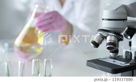 Microscope is near a scientist, wearing a lab coat and pink protective gloves, and holding beaker with a yellow oily liquid in laboratory setting, close up. Medicine and science Microscope is near a scientist, wearing a lab coat and pink protective gloves, and holding beaker with a yellow oily liquid in laboratory setting, close up. Medicine and science 119337880