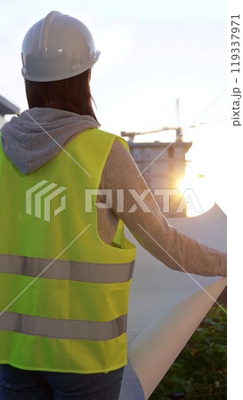 Woman an architect or engineer wearing safety hard hat and vest holding blueprint while inspecting a construction site at sunrise, vertical back view. Architecture and engineering Woman an architect or engineer wearing safety hard hat and vest holding blueprint while inspecting a construction site at sunrise, vertical back view. Architecture and engineering 119337971