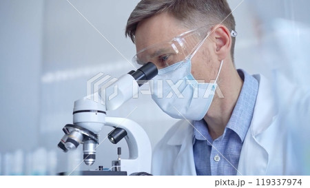 Man scientist with protective glasses, mask and gloves is using microscope in laboratory, portrait of researcher. Microbiology science concept Man scientist with protective glasses, mask and gloves is using microscope in laboratory, portrait of researcher. Microbiology science concept 119337974